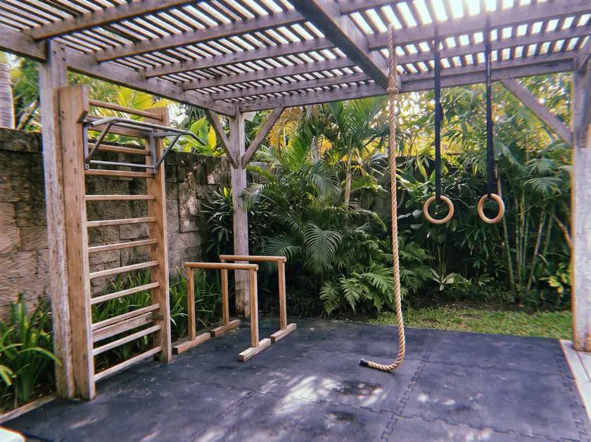 Outdoor gym with wooden stall bars, rings and climbing rope under a pergola